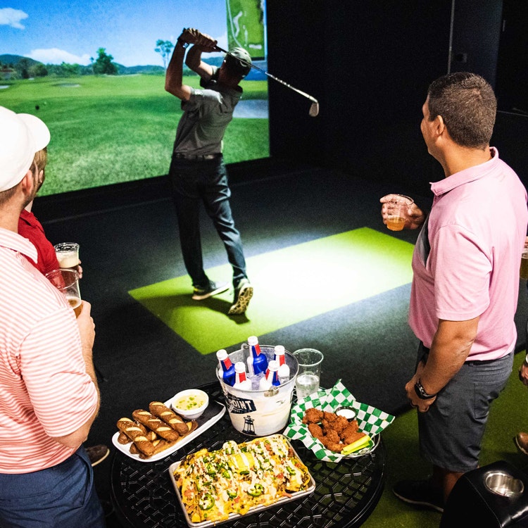people eating and drinking beer around a golf simulator